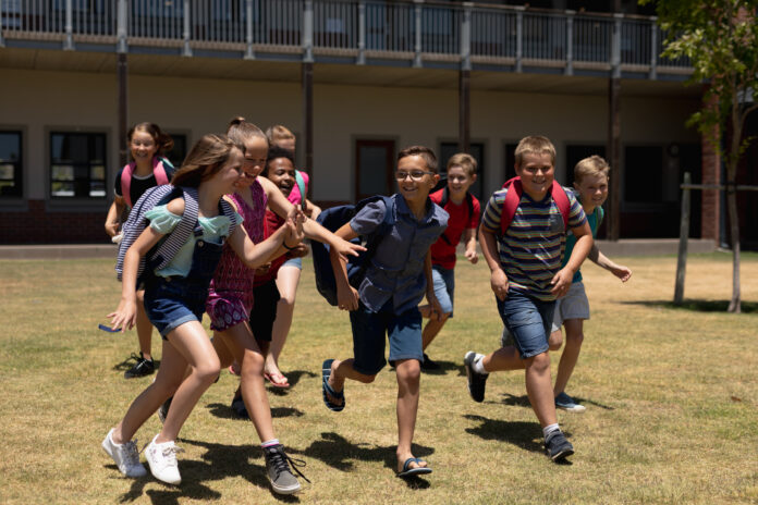 Front view of a diverse group of schoolchildren wearing shorts and rucksacks running in a playing field at elementary school, smiling and laughing